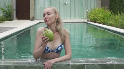 The young slender woman in Bikin drinks coconut milk from a coco on the edge of the pool in the tropical resort