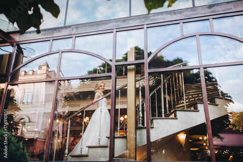 Through the transparent glass of large windows  is visible beautiful woman in a white dress with long train which is climbs up the stairs. Houses and trees are reflected in the windows.