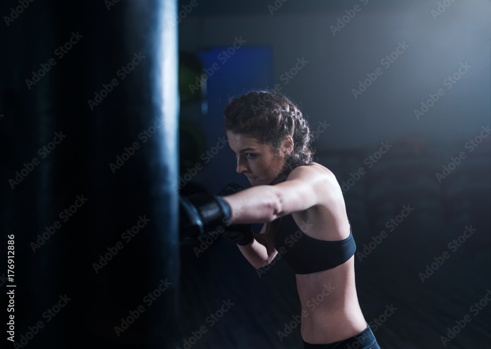 Young fighter boxer fit girl wearing boxing gloves in training with heavy punching bag in gym. Low key image. Woman power