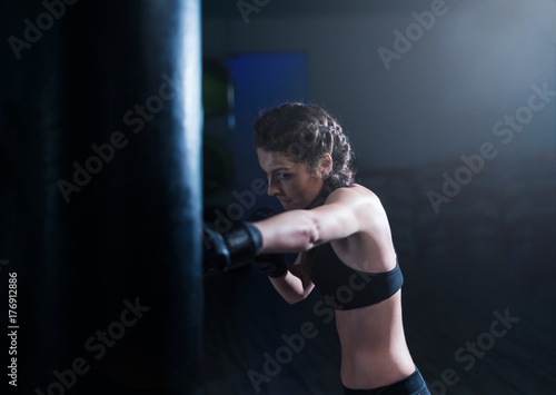 Young fighter boxer fit girl wearing boxing gloves in training with heavy punching bag in gym. Low key image. Woman power