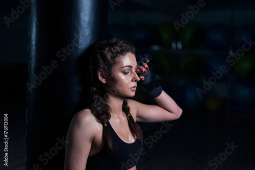 Upset young fighter boxer girl wearing boxing gloves  standing in dark gym near punching bag after loosing fight. She coveres her face with hand.