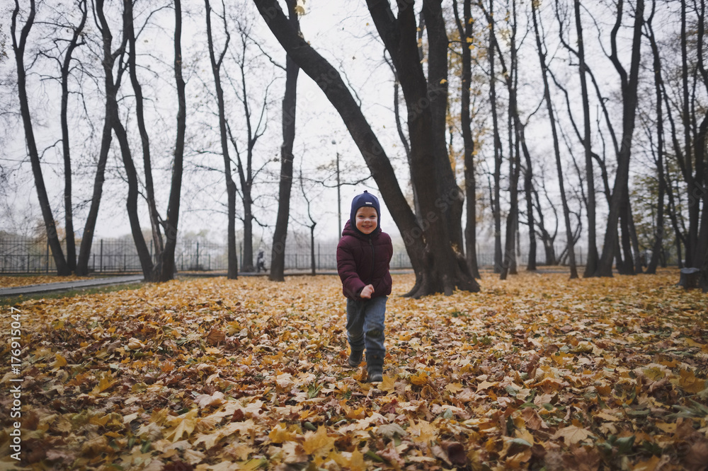 Smiling boy in jacket and hat running in the autumn Park. Around him are yellow fallen leaves.