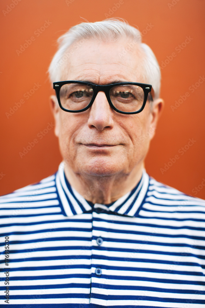 Portrait of an elderly man wearing rimmed glasses standing in front an orange wall. Portrait of an elderly man wearing rimmed glasses standing in front an orange wall.
