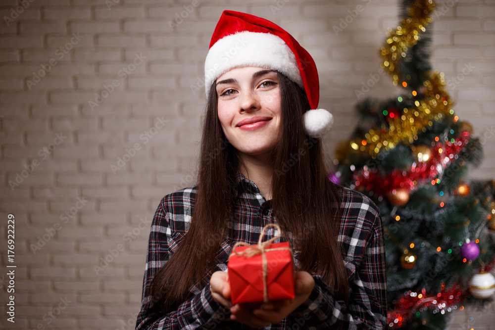 Fototapeta premium Young cute woman in Santa cap holding a gift box on christmas tree background. New Year, Christmas, gift, surprise, emotion, celebration concept