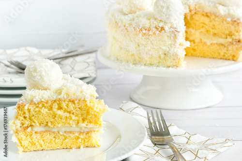 Photo Slice of coconut cake served on white plate, rest of the cake on cake stand