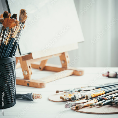 Artistic equipment in painter studio: easel, paint brushes, tubes of paint, palette and paintings on work table of artist.