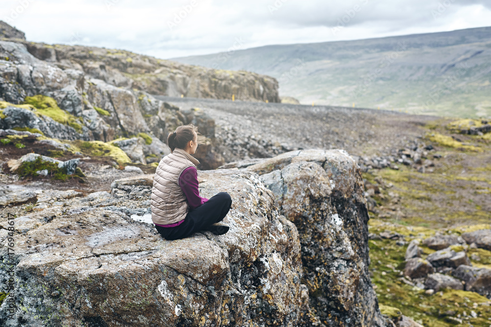 Girl in warm clothing sits on the cliff on background of mountains of Iceland.