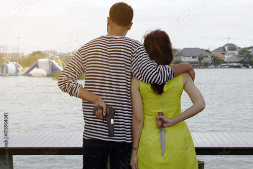 Cheating couple, a man put his arm around woman shoulder looking at the lake in the park. Boyfriend holding a gun girlfriend holding a knife in behind