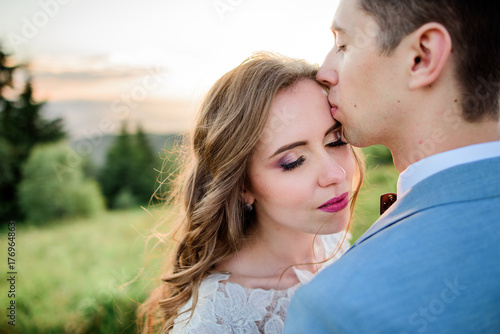 Groom kisses bride's forehead standing on a green hill © IVASHstudio