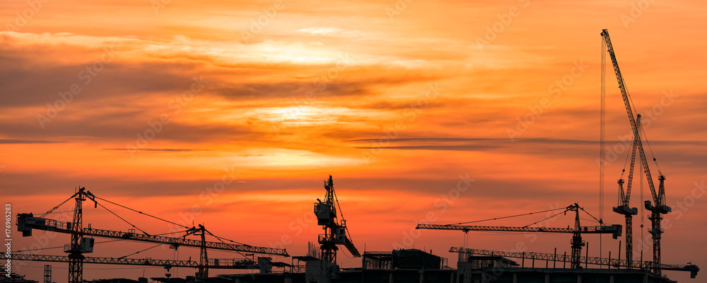 The silhouette image of tower cranes at the real estate and building construction site with the background of golden sky of sun set.
