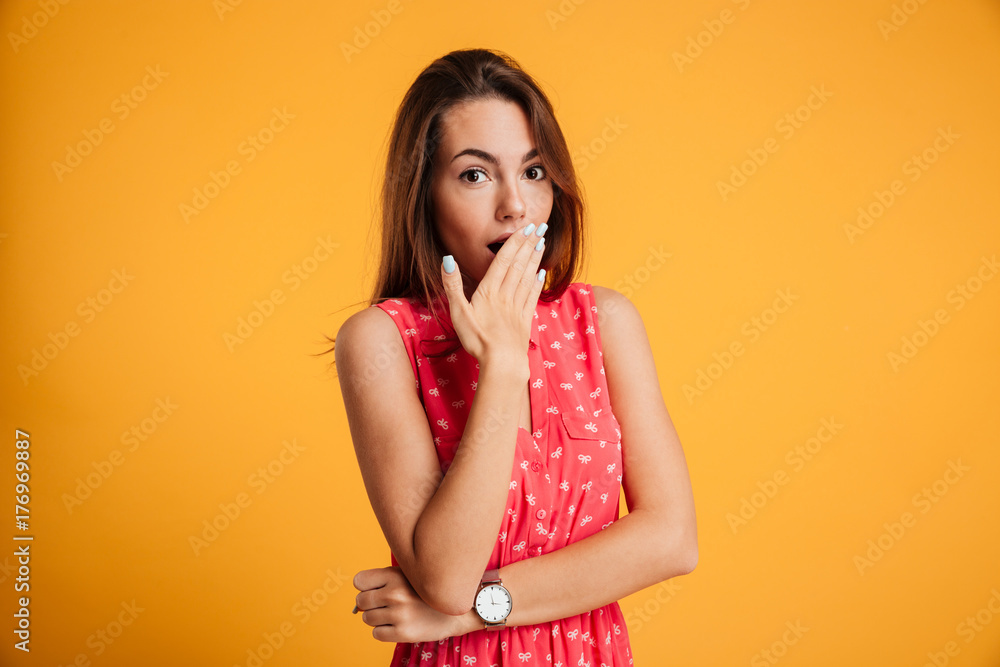 Photo of surprised pretty girl in red dress standing with her hand covering mouth and looking at camera Photo of surprised pretty girl in red dress standing with her hand covering mouth and looking at camera
