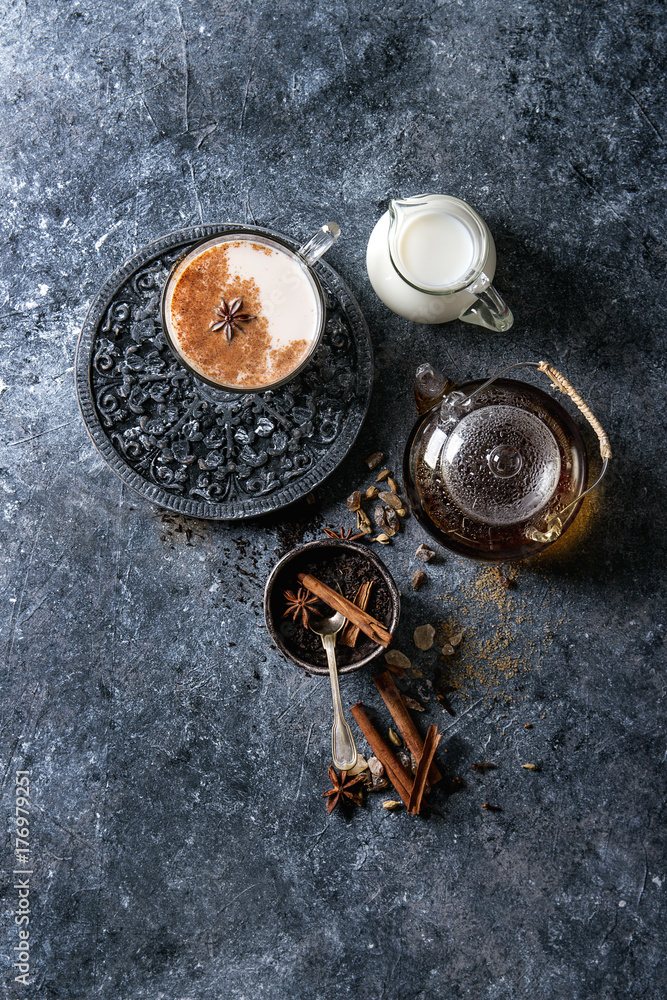Glass cup of traditional indian masala chai tea with ingredients above. Cinnamon, cardamom, anise, sugar, black tea in glass teapot, jug of milk over dark texture background. Top view with space Glass cup of traditional indian masala chai tea with ingredients above. Cinnamon, cardamom, anise, sugar, black tea in glass teapot, jug of milk over dark texture background. Top view with space
