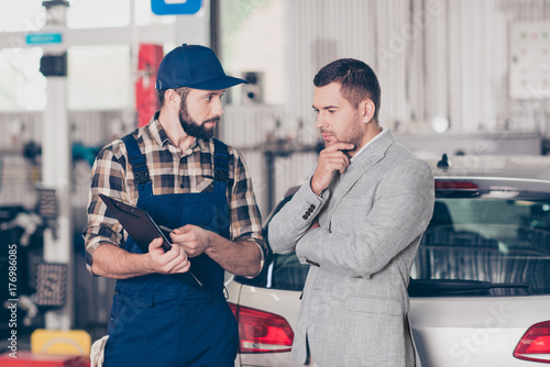 Experienced brunet bearded expert specialist in blue overall is discussing and demonstrates the demages of businessman`s car, he is ponder pensive, focused and concentrated, decisions, options photo