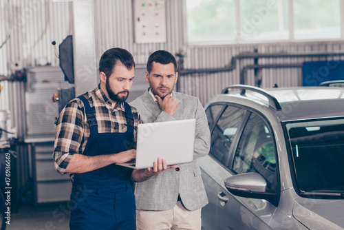 Experienced brunet bearded expert specialist in blue overall and checkered shirt is demonstrating the demages of businessman`s car on his device, he is ponder pensive, focused, decisions, options photo