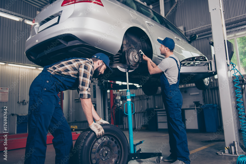 Φωτογραφία Busy colleagues professionals in special blue safety overalls uniform, head wear are torquing lugs nuts of wheel and rolling new rubber tyre, transportation is above
