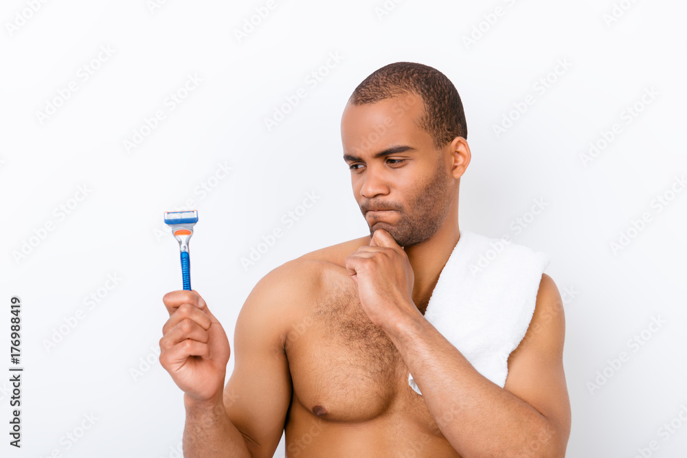 Fototapeta premium Skeptic, unsure, uncertain, doubts concept. Young african nude athletic guy is looking sceptical at razor, has a grimace of distrust on white background