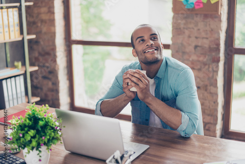 Young dreaming american worker is thinking in front of laptop at work place. He is happy, smiling, behind him is window and brick wall photo