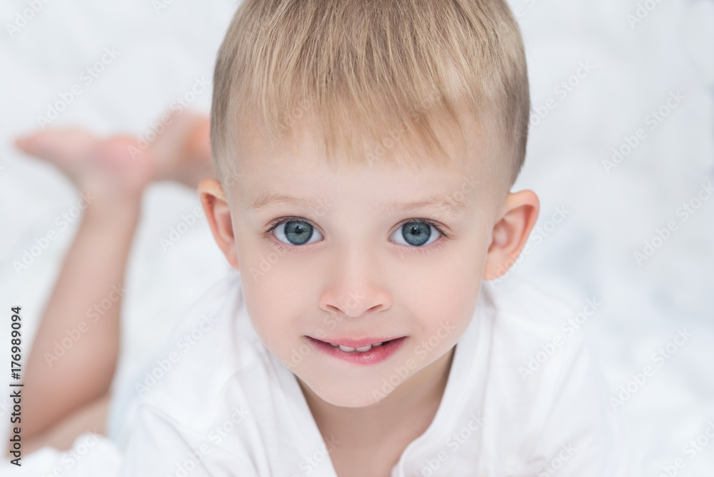 Closeup of smiling little boy lying on white blanket on the bed. Happy cute child with big blue green eyes looking at the camera