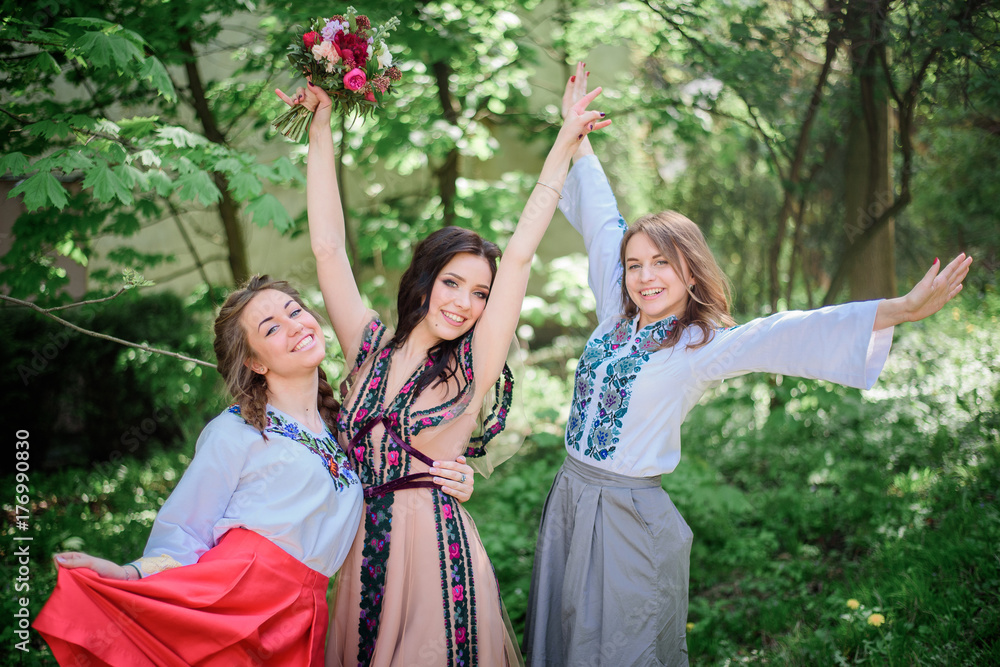 Fototapeta premium Pretty bride and her bridesmaids in embroidered dresses stand in green park