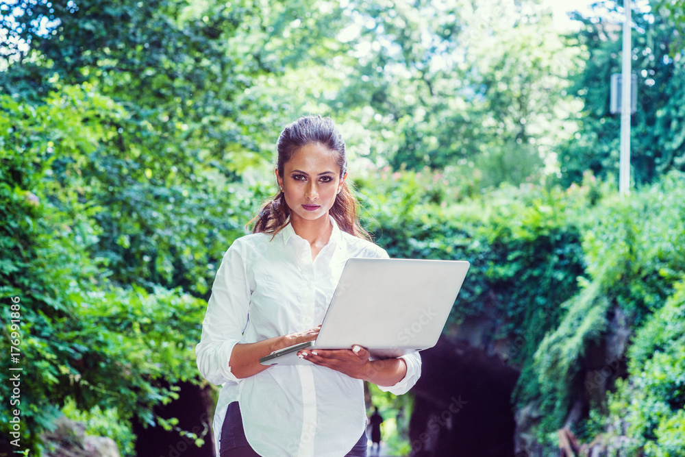 Young Beautiful East Indian American Woman traveling, working in New York, wearing white shirt, holding laptop computer, walking at Central Park. Street bridge with green leaves on background..