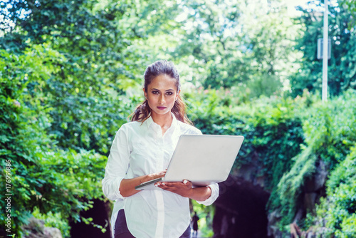 Obraz na płótnie Young Beautiful East Indian American Woman traveling, working in New York, wearing white shirt, holding laptop computer, walking at Central Park. Street bridge with green leaves on background.. w salonie