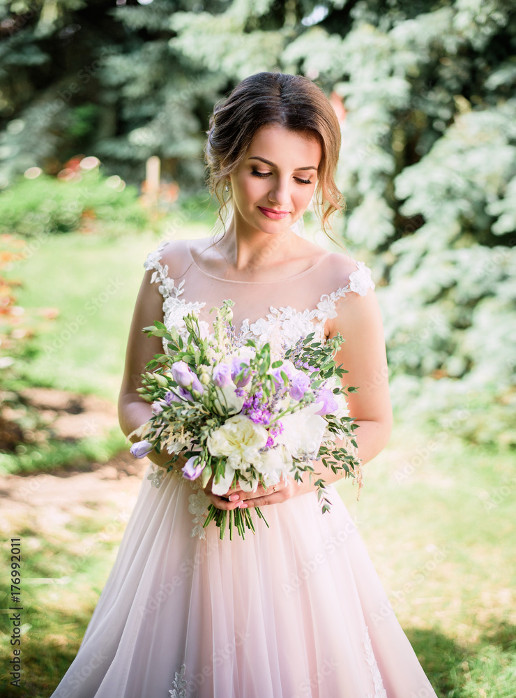 Beautiful bride stands with rich white and violet bouquet in green park