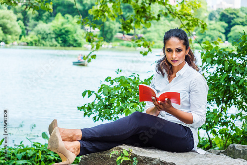 Young East Indian American Woman reading book, relaxing at Central Park, New York, wearing white shirt, black pants, high heels, sitting on rocks by lake, holding red book, looking up, thinking..