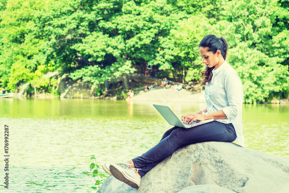 Young East Indian American Woman wearing white shirt, black pants, white sneakers, crossing legs, sitting on rocks by lake at Central Park, New York, looking down, typing, working on laptop computer.
