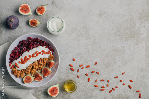 Cereal bowl on minimal concrete table top. Healthy breakfast with granola, goji berry, cranberries, pecan nuts, figs, honey and yogurt. Top view. Copy space..