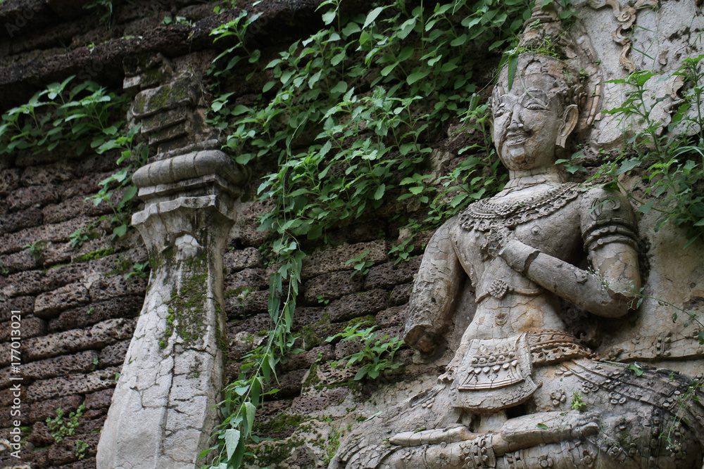 400 years old ruined ancient standing and praying of male angel statue at Chiangmai covered with green leaf, Thailand, buddha statue without some of body part, historical decorated wall temple
