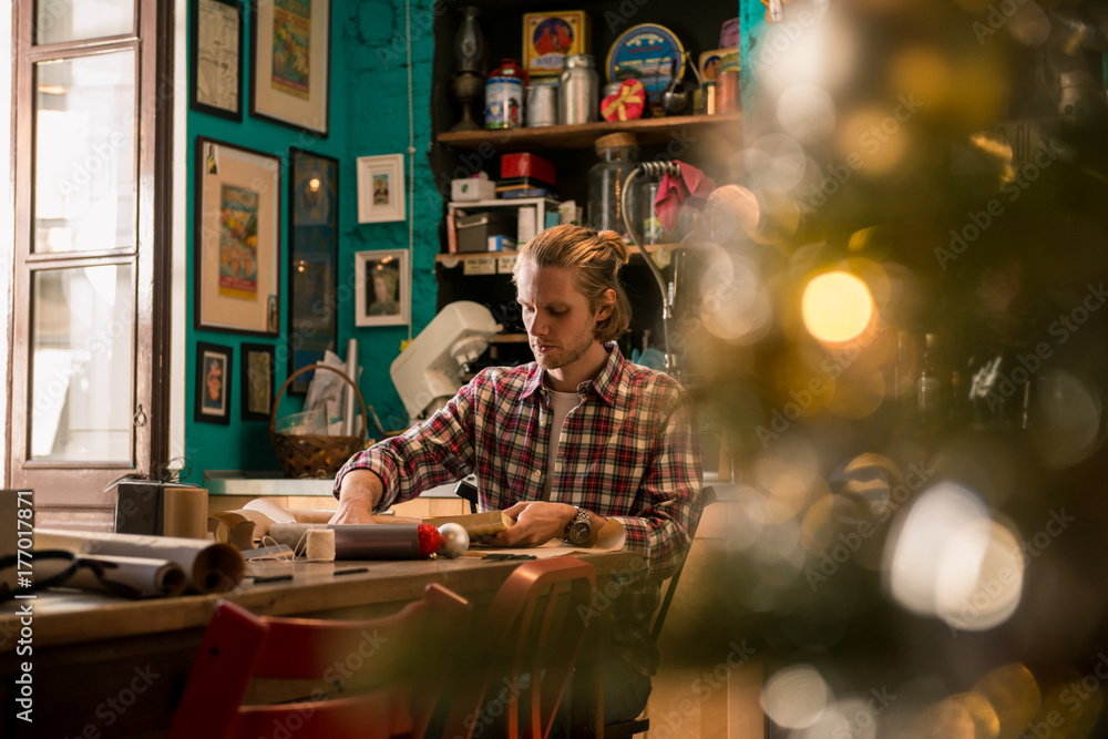 Young adult man with long blonde hair wrapping christmas present with red ribbon seated on wooden table in cozy apartment indoor.