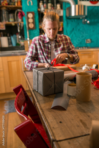 Young adult man with long blonde hair wrapping christmas present with red ribbon seated on wooden table in cozy apartment indoor.