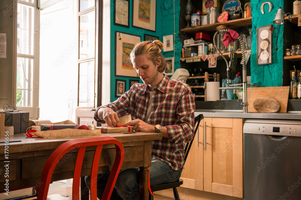 Young adult man with long blonde hair wrapping christmas present with red ribbon seated on wooden table in cozy apartment indoor. Young adult man with long blonde hair wrapping christmas present with red ribbon seated on wooden table in cozy apartment indoor.