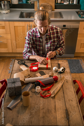 Young adult man with long blonde hair wrapping christmas present with red ribbon seated on wooden table in cozy apartment indoor.