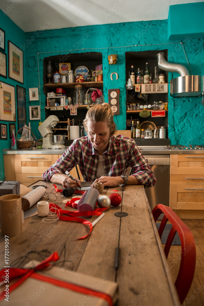Young adult man with long blonde hair writing christmas card seated on wooden table in cozy apartment indoor.