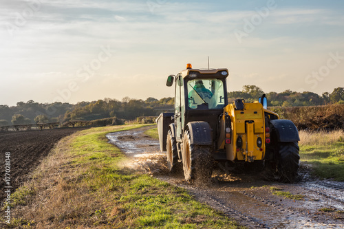 Tractor driving along a wet farm track with a ploughed field to the left.