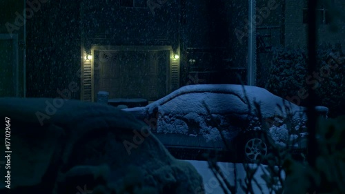 Night time snowfall in a city on peaceful, calm, tranquil evening during winter holidays season on streets of residential suburbs with houses, lights and cars covered with snow by thick snowflakes