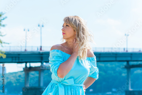 A middle-aged woman, plus in a light blue dress, rests on a city waterfront on a day off. The concept of femininity and romance in the city bustle