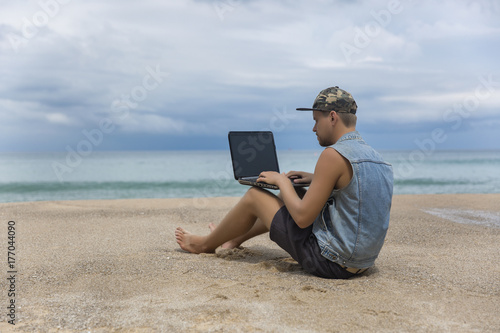 Young man working on laptop while sitting on the beach. Freelance working, social networks concept