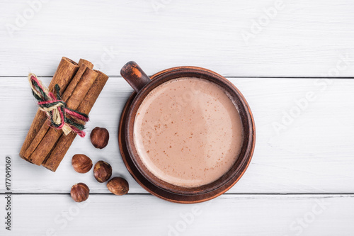 Hot cocoa with milk in brown clay cup, hazelnut and cinnamon sticks on table of white wooden planks, top view