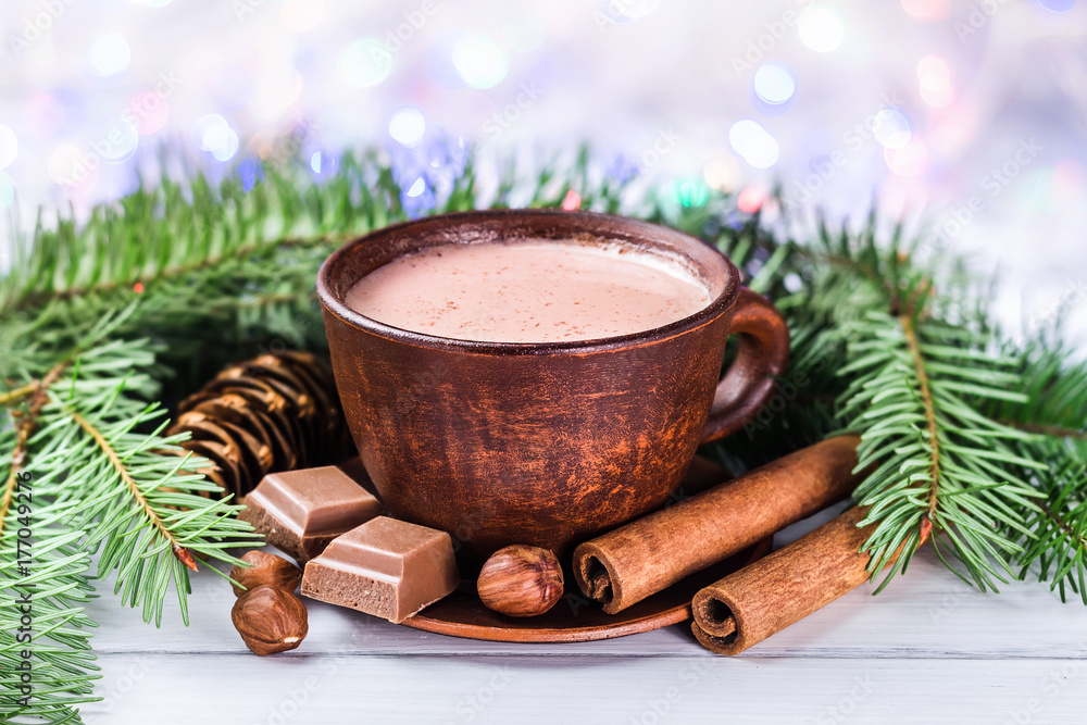 Blurred Christmas lights, cup of hot cocoa with milk, broken chocolate cubes, hazelnut and cinnamon sticks, christmas tree branches, pine cone on white wooden planks Blurred Christmas lights, cup of hot cocoa with milk, broken chocolate cubes, hazelnut and cinnamon sticks, christmas tree branches, pine cone on white wooden planks