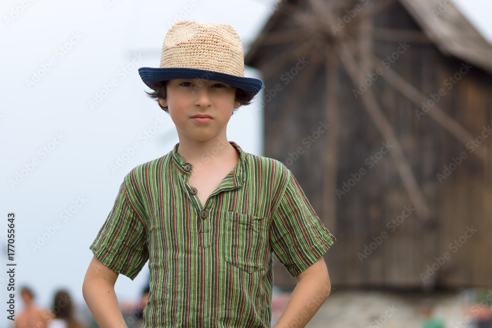 Portrait of a beautiful boy in a short shirt and hat on a blurred background of a wooden windmill.