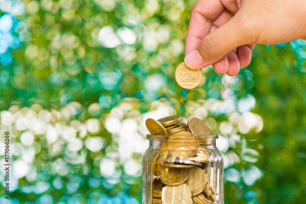 Woman hand put gold coin money in the glass jar on table in garden with green background, for saving banking finance concept. Woman hand put gold coin money in the glass jar on table in garden with green background, for saving banking finance concept.