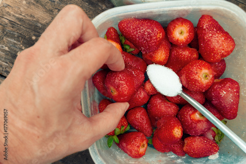 Close-up macro of man's hand taking a strawberry from a pile with metal teaspoon full of sugar