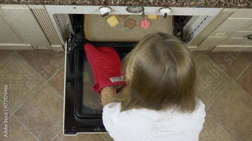 Top view of young white blonde girl pushing tray with ginger cookies bisquits to oven at home kitchen. High angle top view. CU. Red mitten mitt glves. photo