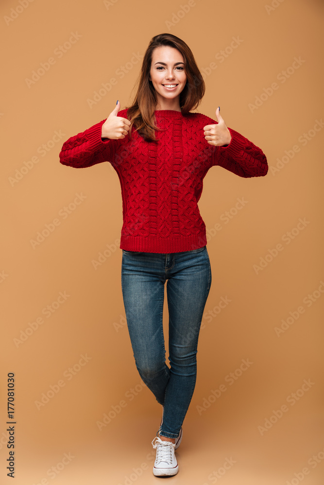 Full length portrait of smiling brunette woman in red knitted sweater and jeans showing thumbs up gesture with two hands Full length portrait of smiling brunette woman in red knitted sweater and jeans showing thumbs up gesture with two hands