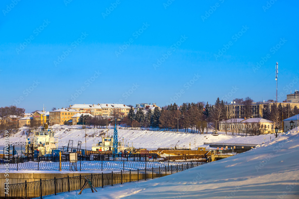 The embankment with a metal fence is covered with snow, barges are at the pier. Urban harbor in winter Uglich, Russia.