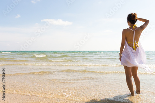 Young lady look towards the ocean in white dress at the beach. Summer vacation and travel concept. Traveling to tropical areas.