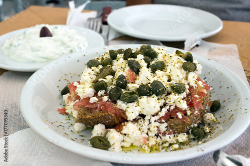 Dakos greek appetizer, dried bread, tomatoes, feta, or mizithra cheese, capers, oregano; Tzatziki in background. Taken in Naxos photo