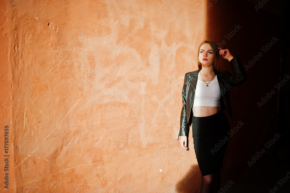 Elegant blonde girl wear on black leather jacket posing at streets of town background old wall with shadows.